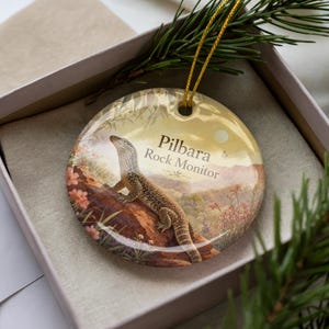 May include: Round ceramic ornament featuring a Pilbara Rock Monitor lizard illustration. The ornament has a gold string for hanging and is presented in a gift box with greenery. The background depicts a desert landscape.