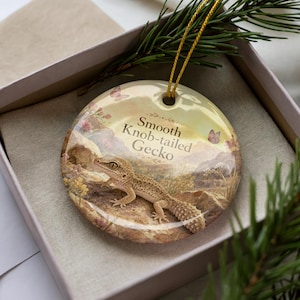 May include: Round ceramic ornament featuring a Smooth Knob-tailed Gecko. The ornament has a desert landscape design with a gecko, butterflies, and floral accents. It includes a gold string for hanging. The ornament is presented in a gift box.