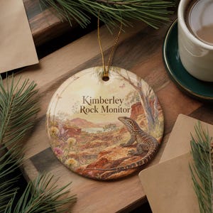 May include: A round ceramic ornament featuring a Kimberley Rock Monitor lizard in a natural landscape. The ornament has a gold string for hanging and includes the text "Kimberley Rock Monitor". The background depicts a scenic view with a lake and trees.
