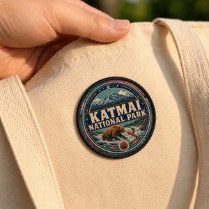 May include: A circular embroidered patch featuring a brown bear catching a salmon in a river with mountains in the background. The text KATMAI NATIONAL PARK is printed in the center. The patch is attached to a cream-colored canvas tote bag.