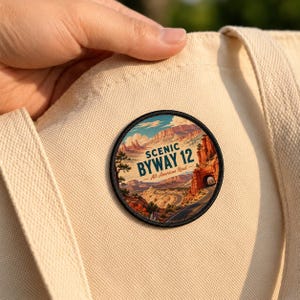 May include: A circular embroidered patch featuring a landscape illustration of a winding road through red rock canyons. The text reads SCENIC BYWAY 12 All-American Road. The patch is attached to a light beige canvas tote bag with a black stitched border.
