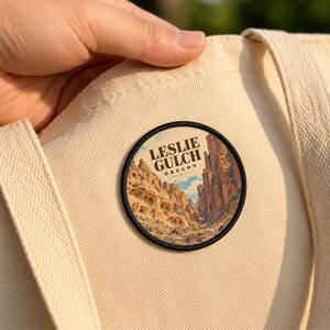 May include: A circular embroidered patch featuring a landscape illustration of Leslie Gulch, Oregon, with rocky canyon walls and desert vegetation. The text LESLIE GULCH OREGON is printed in dark brown letters on a cream-colored canvas tote bag.