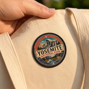 May include: A circular embroidered patch featuring a landscape illustration of Yosemite National Park with mountains, a waterfall, and a road. The text YOSEMITE NATIONAL PARK is printed in blue on a cream background, all on a beige canvas tote bag.