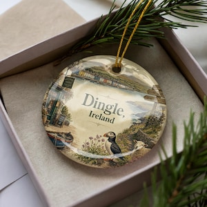 May include: Round ceramic ornament featuring a scenic view of Dingle, Ireland, with a puffin and sheep. The ornament has a gold string for hanging and is presented in a gift box. The text "Dingle, Ireland" is prominently displayed.