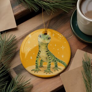 May include: A round, glossy yellow ceramic ornament featuring a detailed illustration of a green lizard with dark spots. The lizard stands on a light-colored patch, surrounded by white line-drawn stars and suns. A gold string is attached to the top.