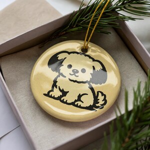May include: A round, cream-colored ceramic ornament featuring a black illustration of a cartoon dog. The ornament has a gold string for hanging and is presented in a gift box with a sprig of evergreen.