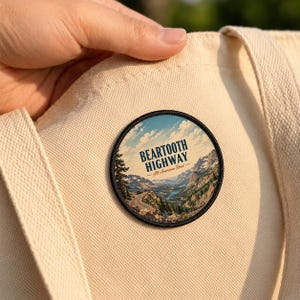 May include: A circular embroidered patch attached to a beige canvas tote bag. The patch features a scenic mountain landscape with a winding road, blue sky, and the text BEARTOOTH HIGHWAY All-American Road in dark blue and red lettering.