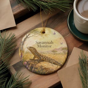 May include: Round ceramic ornament featuring a Savannah Monitor lizard in a natural landscape. The ornament has a gold string for hanging and includes the text "Savannah Monitor". The background shows a savanna scene with trees and butterflies.