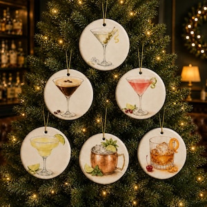 May include: A set of six round, white ceramic ornaments hanging on a Christmas tree. Each ornament features a detailed illustration of a cocktail: a martini with olives, an espresso martini, a cosmopolitan, a margarita, a Moscow mule, and an old fashioned.