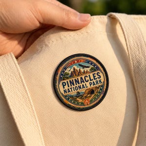 May include: A circular embroidered patch featuring a landscape illustration of Pinnacles National Park with a flying bird, rock formations, and trees. The text reads PINNACLES NATIONAL PARK in bold blue letters on a cream background.