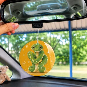 May include: A round car air freshener hanging from a rearview mirror, featuring a green snake illustration on a yellow background with small white star accents. The circular ornament is suspended by a thin white string in a vehicle interior.