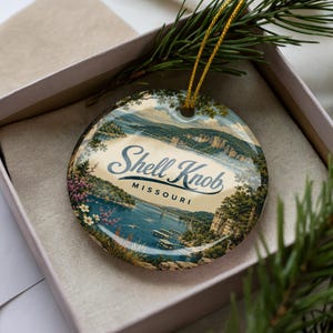 May include: A round ceramic ornament featuring a scenic illustration of Shell Knob, Missouri, with a lake, bridge, and surrounding trees. The text Shell Knob Missouri is printed in the center. A gold string is attached for hanging purposes.