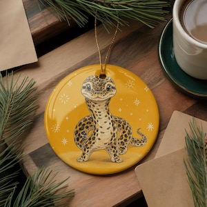 May include: A round, yellow ceramic ornament featuring a detailed illustration of a spotted leopard gecko. The gecko has a cream-colored underbelly and dark brown spots. The background includes small white star and sun icons and a gold string.