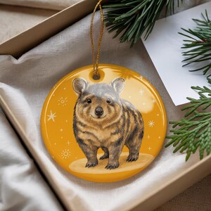 May include: A round, glossy, mustard-yellow ceramic ornament featuring a detailed illustration of a brown, furry quokka standing on a light tan circular patch. The background is decorated with small white star and sunburst icons and includes a gold string.