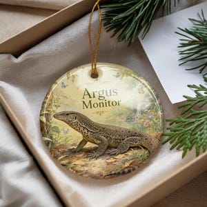 May include: Round ceramic ornament featuring an Argus Monitor lizard illustration. The ornament has a gold string for hanging. The background shows a natural scene with butterflies and the text "Argus Monitor".
