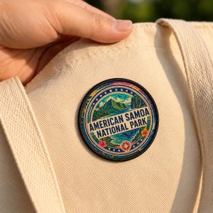 May include: A circular embroidered patch featuring a landscape of American Samoa National Park, with green mountains, blue water, and tropical flowers. The text reads AMERICAN SAMOA NATIONAL PARK in dark blue letters on a cream canvas tote bag.