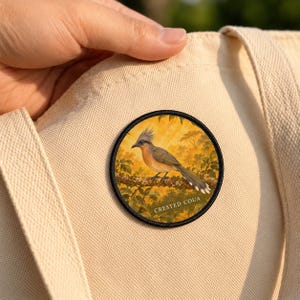 May include: A circular embroidered patch featuring a Crested Coua bird perched on a branch against a golden yellow forest background. The patch has a black stitched border and is attached to a light beige canvas tote bag fabric.