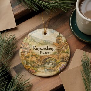 May include: Round ceramic ornament featuring a scenic watercolor painting of Kaysersberg, France, with a bridge, buildings, and a river. The ornament has a gold string for hanging and the text "Kaysersberg, -France-".