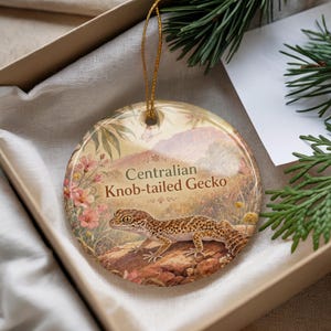 May include: A round ceramic ornament featuring a Centralian Knob-tailed Gecko illustration. The ornament has a gold string for hanging and is presented in a gift box. The background depicts a landscape with flowers and mountains.