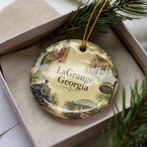 May include: A round Christmas ornament featuring a vintage-style illustration of LaGrange, Georgia. The ornament includes images of a train, buildings, and the text "LaGrange, Georgia". It has a gold string for hanging and is presented in a gift box.