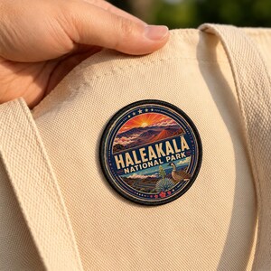 May include: A circular embroidered patch featuring a landscape of Haleakala National Park with a sunrise over volcanic mountains, a nene goose, and the text HALEAKALA NATIONAL PARK, attached to a cream-colored canvas tote bag with visible fabric texture.