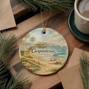 May include: Round ceramic ornament featuring a beach scene with the text "Carpinteria, California." The image depicts a sandy beach, ocean, palm trees, and a sailboat under a blue sky. Includes a gold string for hanging.