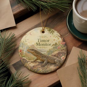 May include: Round ceramic ornament featuring a Timor Monitor lizard in a natural landscape with the text "Timor Monitor". The ornament has a gold string for hanging and is decorated with flowers and butterflies.