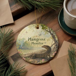 May include: Round ceramic ornament featuring a detailed illustration of a mangrove monitor lizard in a natural habitat. The ornament has the text "Mangrove Monitor" and is decorated with butterflies and flowers. A gold string is attached for hanging.