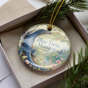 May include: Round ceramic ornament featuring a blue tree monitor lizard illustration. The ornament has a gold string for hanging and includes the text "Blue Tree Monitor". The background shows a forest scene with butterflies and flowers.