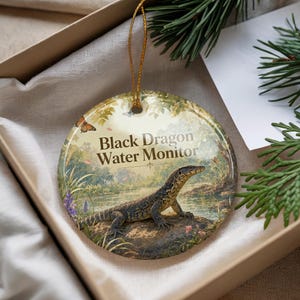 May include: Round ceramic ornament featuring a Black Dragon Water Monitor lizard in a naturalistic setting. The ornament has a gold string for hanging and is presented in a gift box. The text "Black Dragon Water Monitor" is printed on the front.