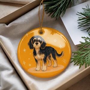 May include: A round, yellow ceramic ornament featuring a black and tan dog illustration. The dog is standing on a light brown oval, with white star and sunburst designs around it. A gold string is attached for hanging.