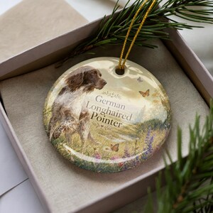 May include: A round ceramic ornament featuring a German Longhaired Pointer in a field setting with butterflies. The ornament has a gold string for hanging and is presented in a gift box.