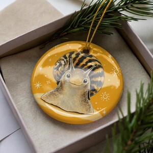 May include: A round, glossy, mustard-yellow ceramic ornament featuring a detailed illustration of a snail with a brown-striped shell. The background includes small white star and sun icons. A thin gold cord is attached for hanging the ornament.