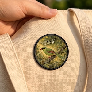 May include: A circular embroidered patch featuring a green magpie perched on a branch with the text COMMON GREEN MAGPIE printed above it. The patch is attached to a light beige canvas fabric with a visible woven texture and stitched seams.