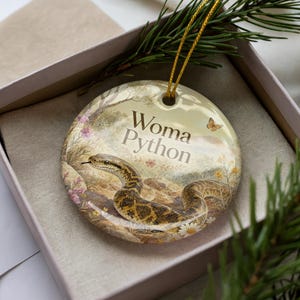 May include: A round ceramic ornament featuring a Woma Python illustration. The ornament has a gold string for hanging and is set in a gift box. The design includes the text "Woma Python" and a butterfly.