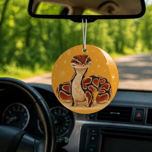 May include: A round car air freshener hanging from a rearview mirror, featuring a mustard yellow background with a brown and cream-colored ball python illustration and small white star accents, suspended by a thin white elastic string.