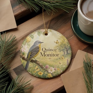May include: Round ceramic ornament featuring a bird perched on a branch, with the text "Quince Monitor." The design includes butterflies, flowers, and a natural landscape. A gold string is attached for hanging.