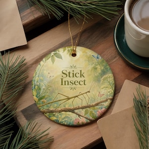 May include: Round ceramic ornament with a nature scene featuring a stick insect on a branch, with the text "Stick Insect". The background includes butterflies, flowers, and foliage. A gold string is attached for hanging.