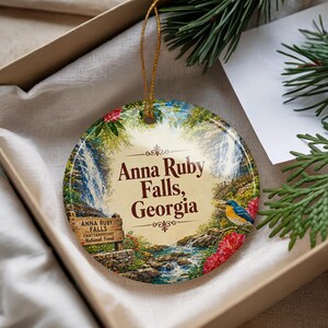 May include: A round ceramic ornament featuring a printed illustration of Anna Ruby Falls in Georgia. The design includes a waterfall, a wooden sign reading Anna Ruby Falls Chattahoochee National Forest, a blue bird, and red flowers.