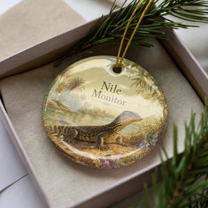 May include: Round ceramic ornament featuring a Nile Monitor lizard in a naturalistic landscape. The ornament has a gold string for hanging and the words "Nile Monitor" are printed on the front. The ornament is presented in a gift box.