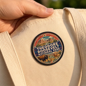 May include: A circular embroidered patch featuring a landscape of Theodore Roosevelt National Park with a bison, horses, and a prairie dog. The text reads THEODORE ROOSEVELT NATIONAL PARK in bold letters, set against a cream-colored canvas tote bag.