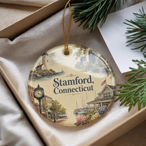 May include: A round ceramic ornament featuring a detailed illustration of Stamford, Connecticut. The design includes a lighthouse, cityscape, harbor scene, and the text "Stamford, Connecticut". The ornament has a gold string for hanging.