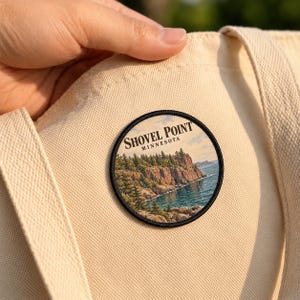 May include: A circular patch featuring a landscape illustration of Shovel Point, Minnesota, with rocky cliffs, evergreen trees, and blue water. The text reads Shovel Point Minnesota. The patch is attached to a cream-colored canvas tote bag.