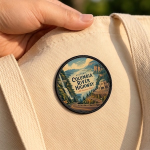 May include: A circular embroidered patch featuring a landscape illustration of the Historic Columbia River Highway, including a waterfall, a winding road, and a stone building, attached to a cream-colored canvas tote bag with a black border.
