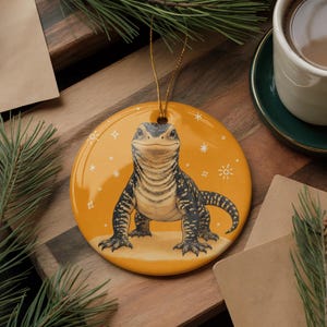 May include: A round, glossy, mustard-yellow ceramic ornament featuring a detailed illustration of a black and tan monitor lizard. The lizard is centered, facing forward, with a light-colored underbelly. White line-art stars and suns surround it.