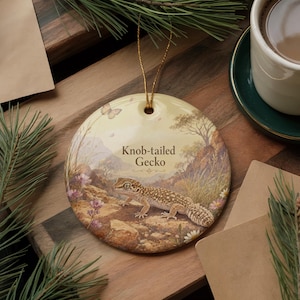 May include: Round ceramic ornament featuring a Knob-tailed Gecko illustration. The ornament has a gold string for hanging. The gecko is depicted in a naturalistic setting with text that reads "Knob-tailed Gecko". The background includes a landscape with rocks and flowers.