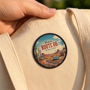 May include: A circular embroidered patch featuring a desert landscape with a light blue vintage car on a road, a tall cactus, and a motel sign. The text reads Historic ROUTE 66 All-American Road. The patch is attached to a beige canvas fabric.