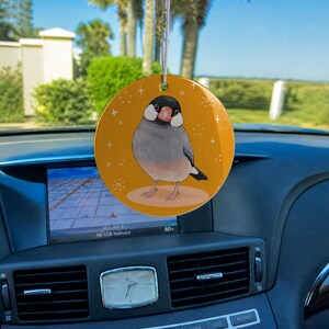 May include: A round, orange car ornament featuring a detailed illustration of a Java sparrow with a black head, white cheek patches, and a grey body. The bird stands on a light tan oval, surrounded by small white star and sparkle accents.
