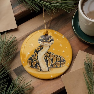 May include: A round, glossy ceramic ornament featuring a detailed illustration of a ball python with brown and black markings against a vibrant mustard yellow background decorated with small white stars and sunburst icons, suspended by a gold cord.