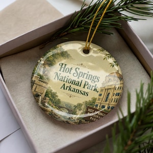 May include: A round ceramic ornament featuring a scenic illustration of Hot Springs National Park, Arkansas. The ornament has a gold string for hanging and is presented in a gift box with a sprig of evergreen.